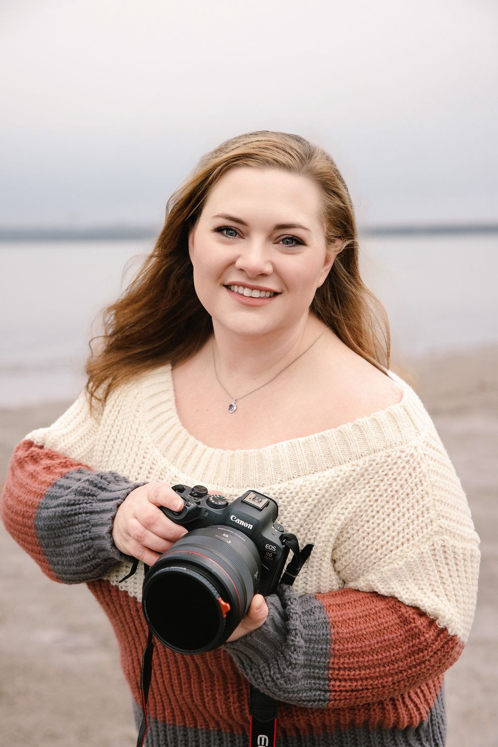 Photographer in cream and rust colored sweater holds camera while smiling on beach during overcast day.