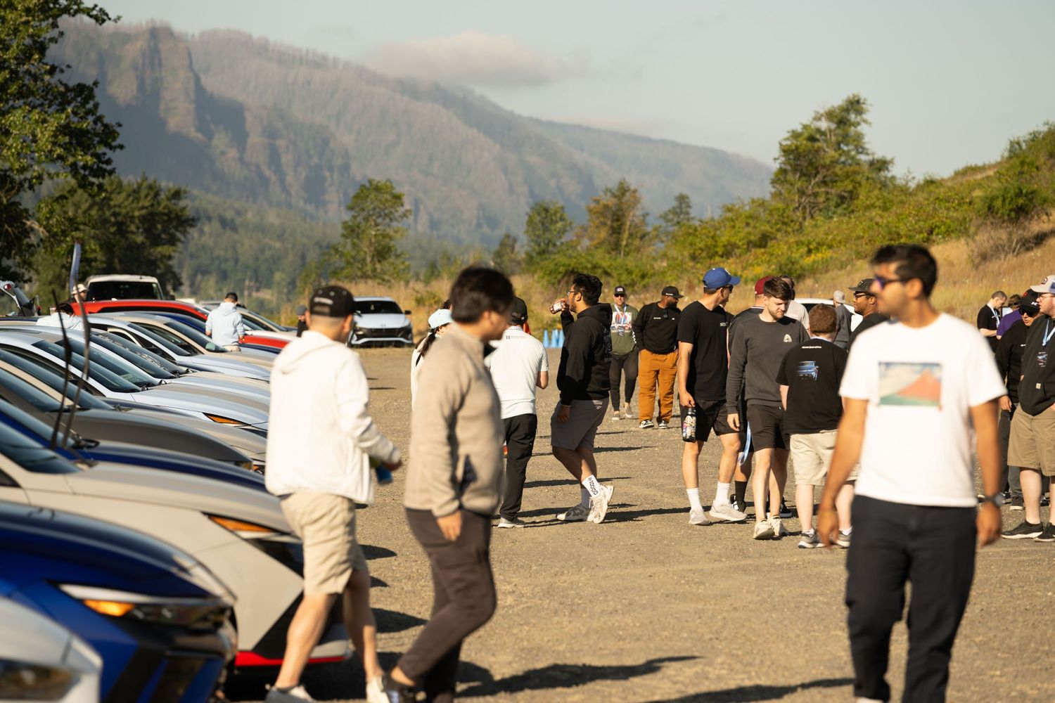 Group of car enthusiasts gather in a parking lot with mountains visible in the background on a sunny day.