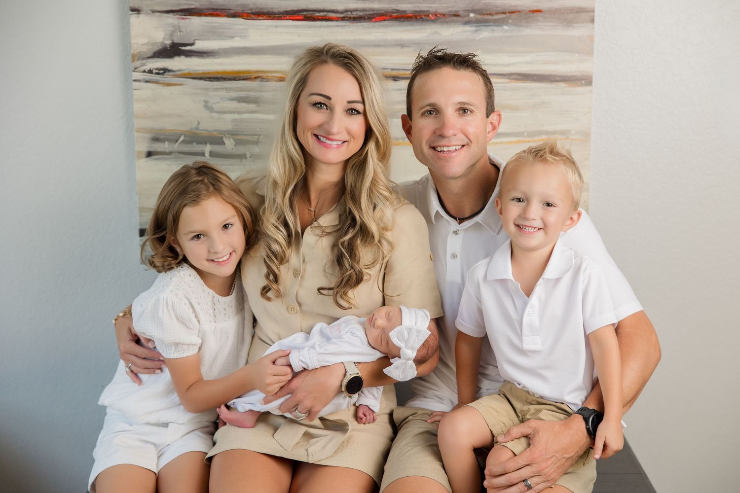 Family wearing coordinated white outfits sits together on grey couch during indoor photo session.