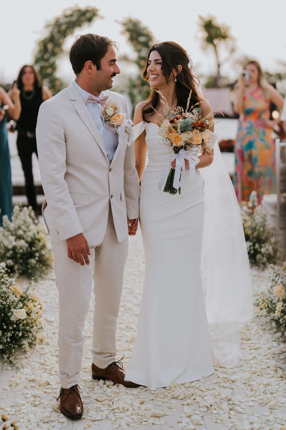 Couple walks along stone path surrounded by white floral arrangements at outdoor wedding.