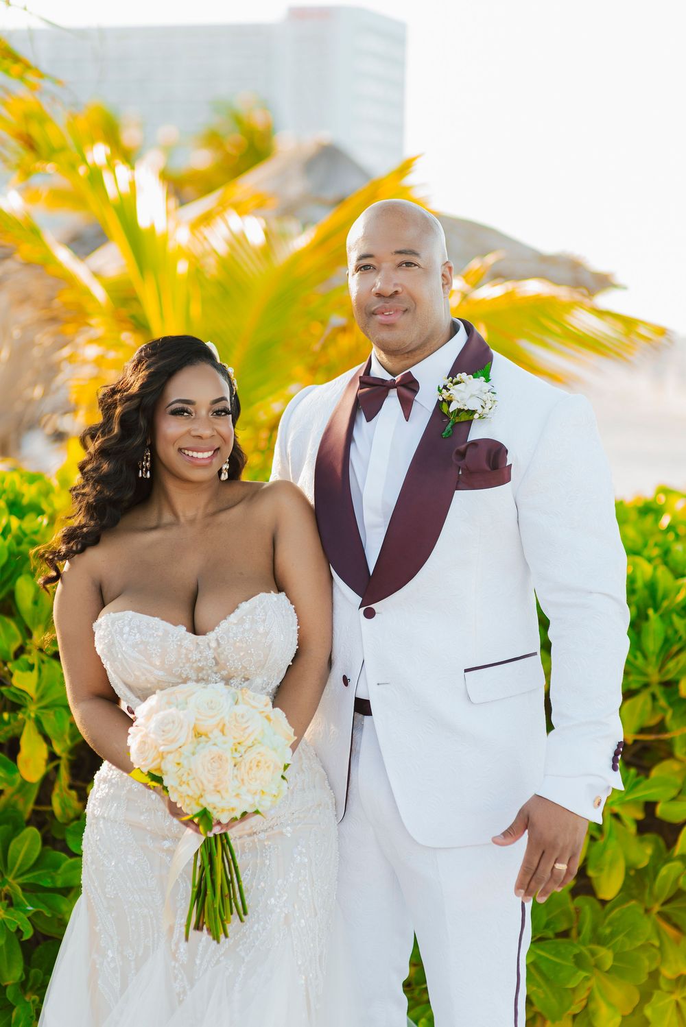 A stunning beach wedding portrait featuring a couple in elegant white and burgundy attire with tropical foliage.