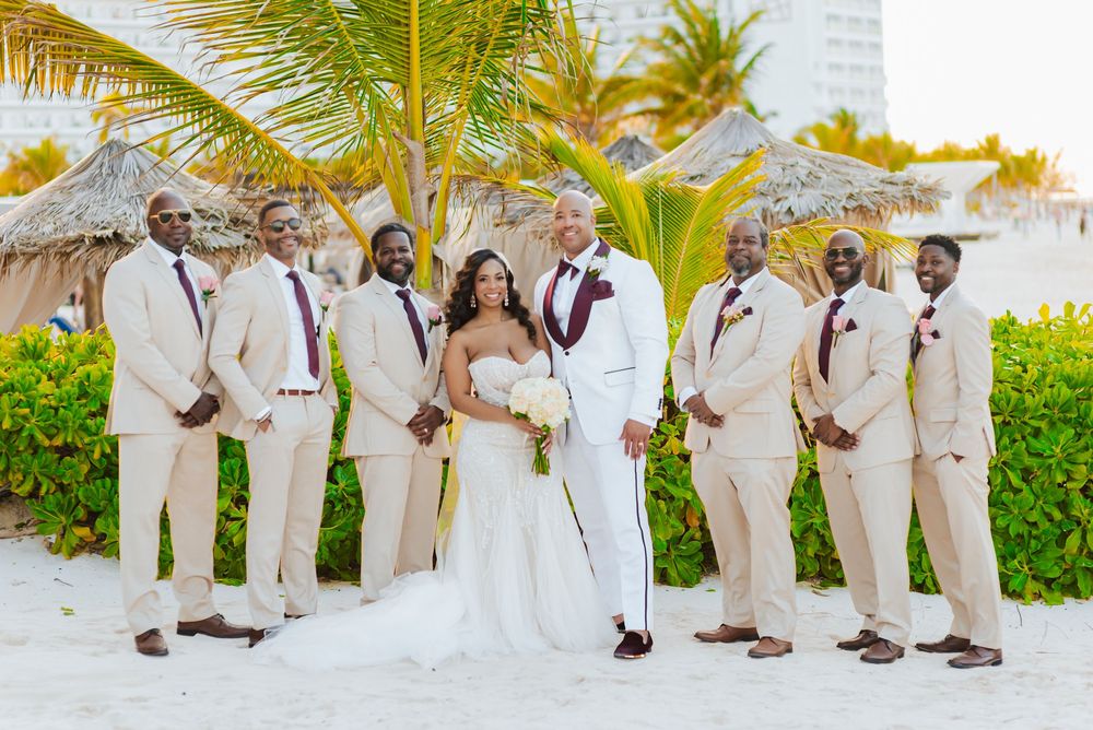 Wedding party poses on beach in coordinated white and beige attire with palm trees in background.