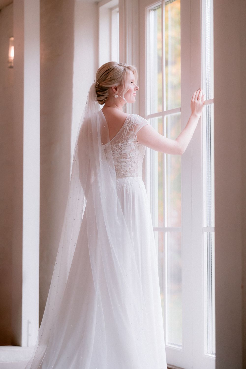 Dreamy sequence of a bride in flowing white dress and veil standing by large windows in natural light.