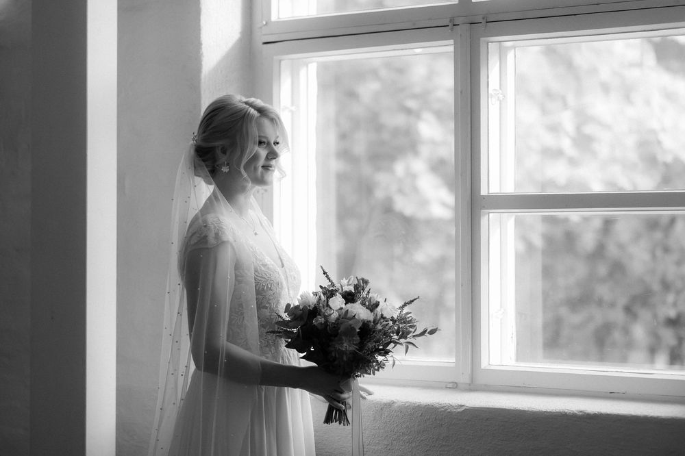 A figure stands in dreamy silhouette by a bright window holding a bouquet, creating an ethereal bridal moment.