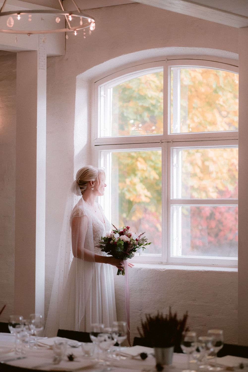 Ethereal bridal portraits in white against arched windows with soft natural light and autumn colors outside.