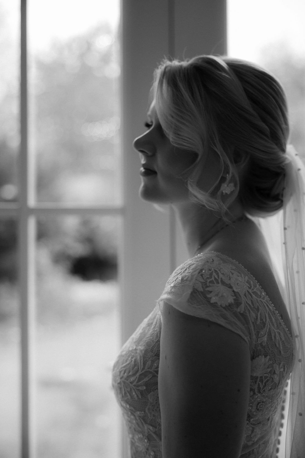 Side view silhouette of bride in lace dress standing by window in soft natural light.