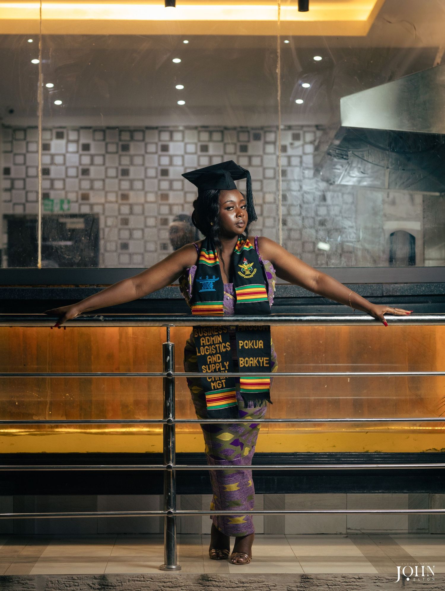 Graduate student in cap and gown poses confidently against modern interior railing with ambient lighting.