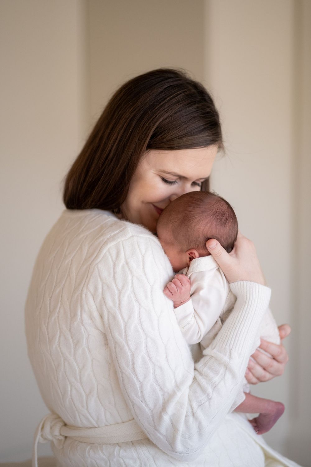 A mother in a white sweater cradles and bonds with her newborn baby in soft natural light.