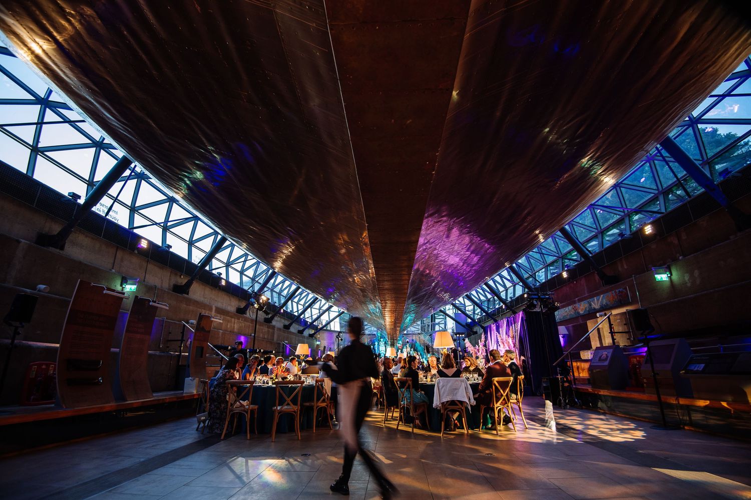 Modern industrial venue with colorful lighting and long dining tables set for an evening event.
