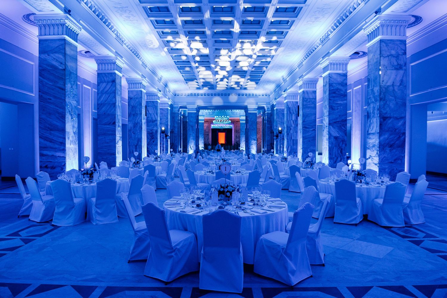 Corporate event space bathed in blue lighting with white-covered chairs and elegant table settings.