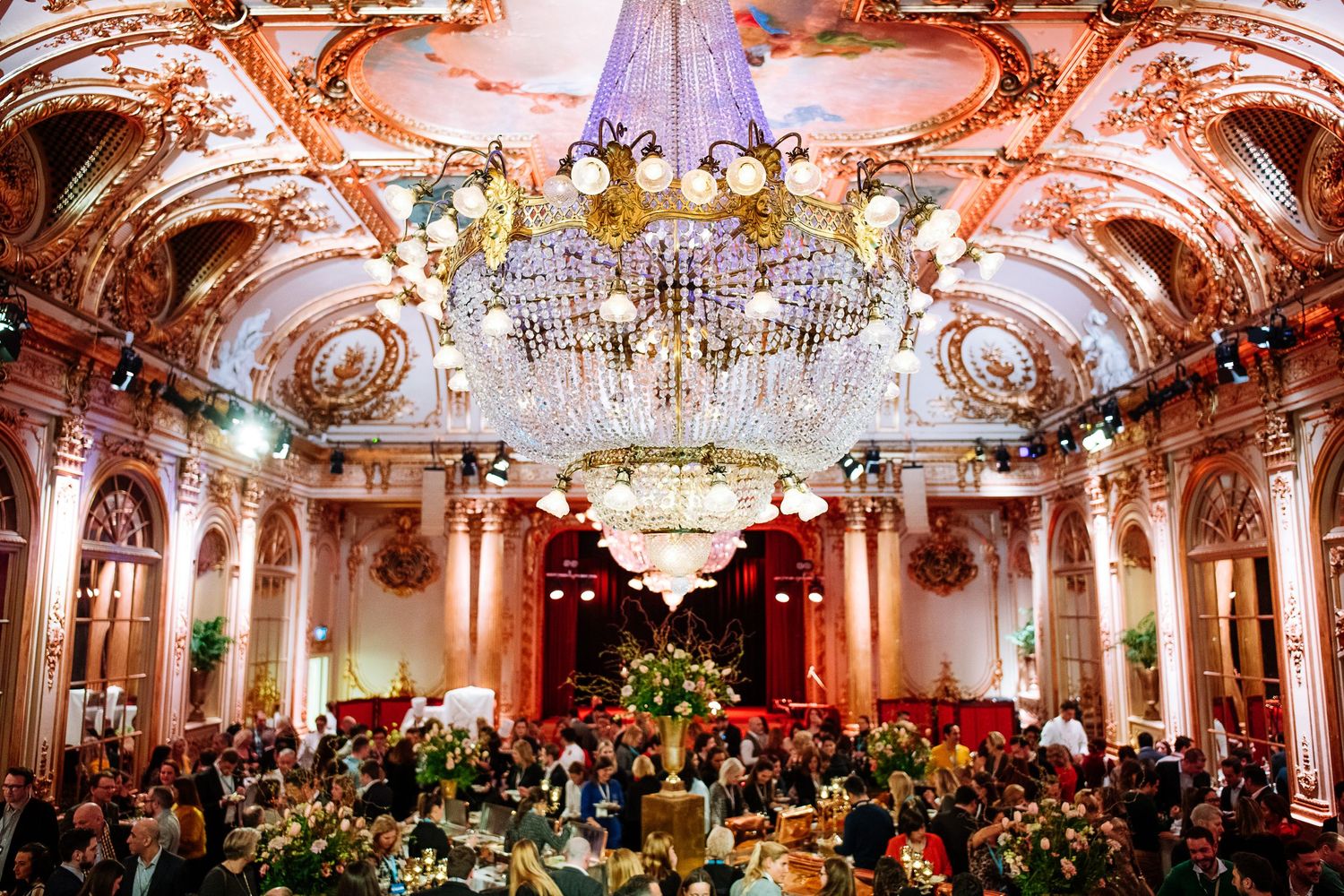 Ornate ballroom with crystal chandeliers and baroque architecture filled with dining tables for an elegant event.