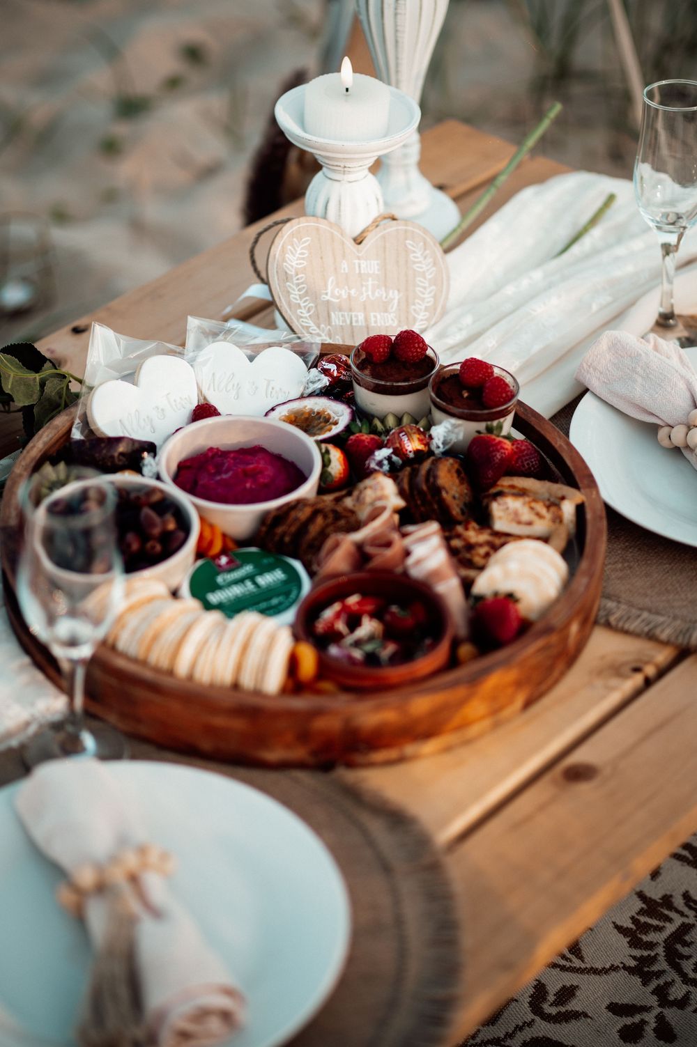 A romantic charcuterie board with wine glasses, candles and red dips on a rustic wooden table setting.