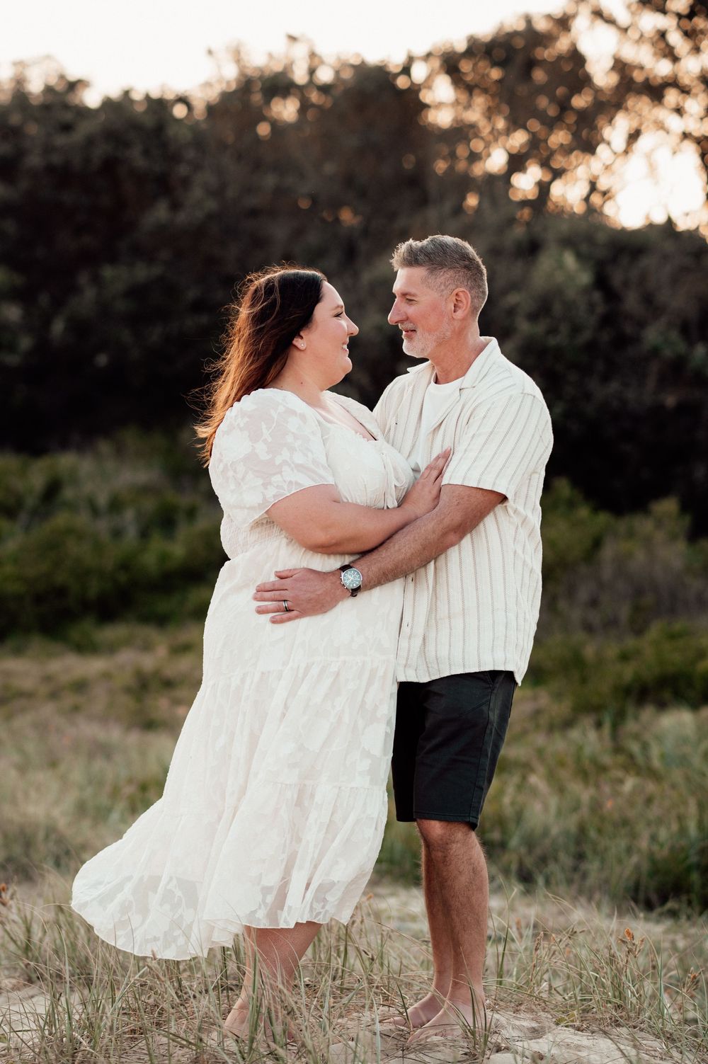 A romantic sunset photoshoot in a grassy field captures a couple in white outfits embracing and dancing together.