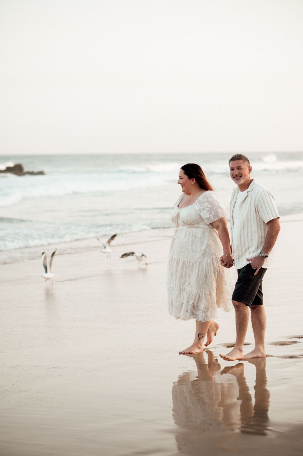 A couple walks barefoot along a beach at sunset while seagulls fly overhead and waves crash in the background.