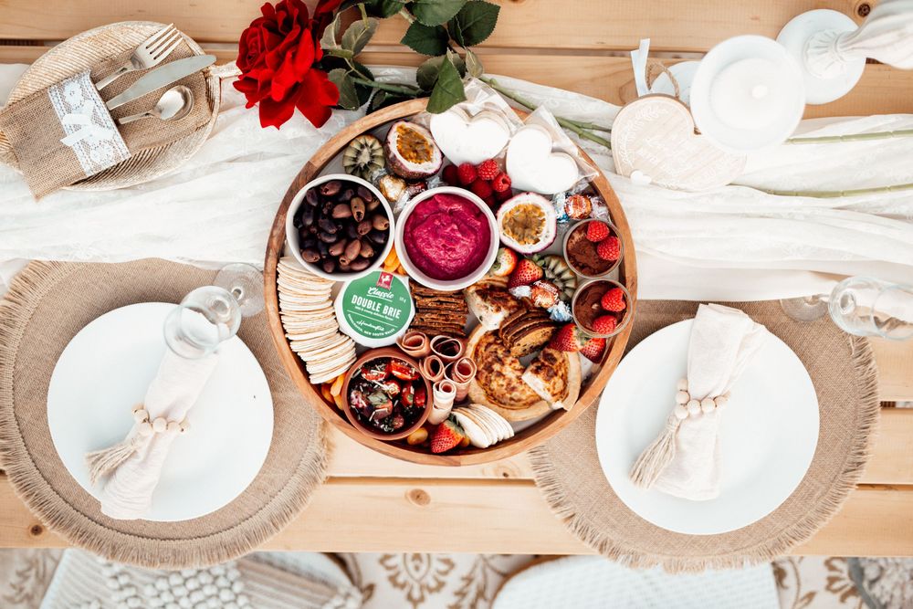 Overhead view of a romantic table setting with charcuterie board, pink dips, white plates and rose decor on wooden surface.
