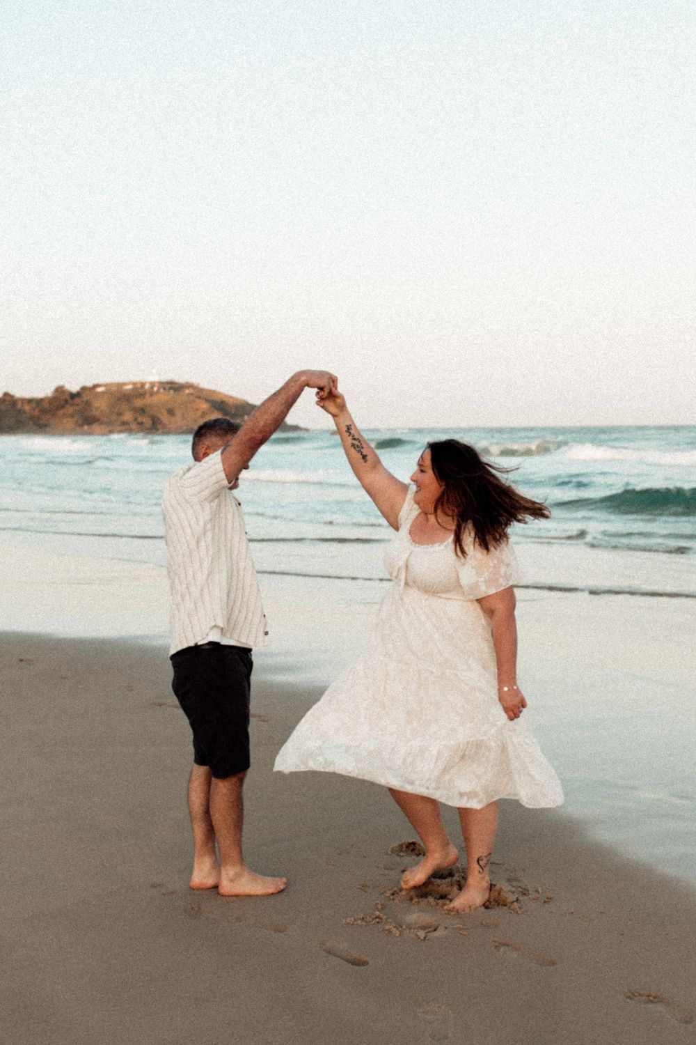 A couple dances together on a beach at sunset, with waves and cliffs in the background.