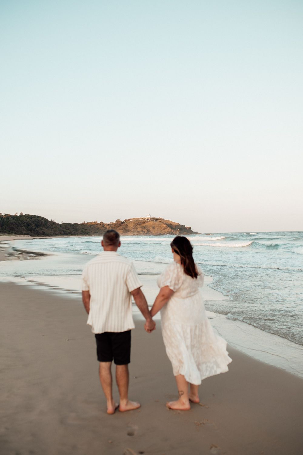 A couple walks hand in hand along a scenic beach with crashing waves and coastal cliffs in the background at sunset.