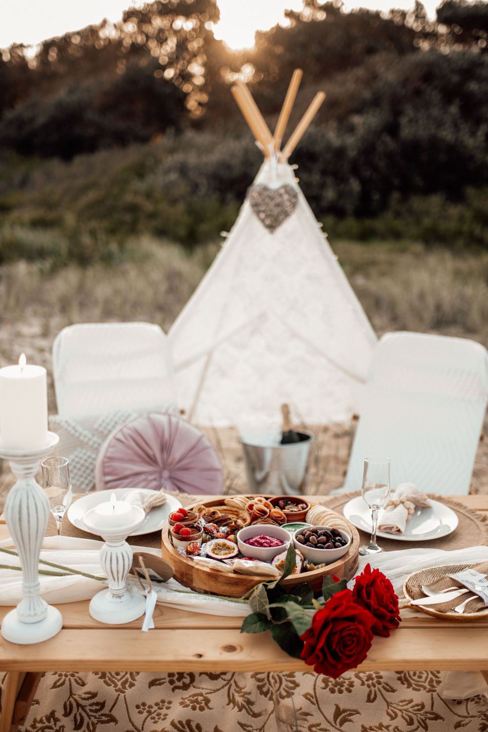 Elegant outdoor table setting with white teepees, wooden table, rose decorations and place settings at sunset.