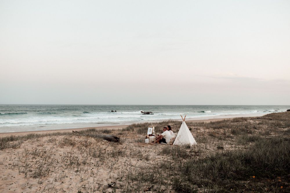Teepee-style tents and campervans dot a scenic coastal landscape with rolling dunes and crashing ocean waves at sunset.