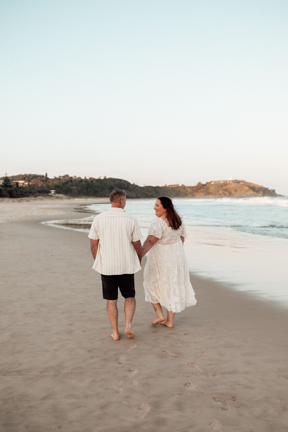 A couple strolls barefoot along a serene beach at sunset with coastal cliffs in the background.