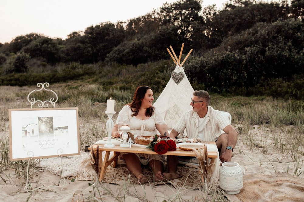 Rustic beach picnic scene with teepee, blankets, and decorative elements at golden hour.