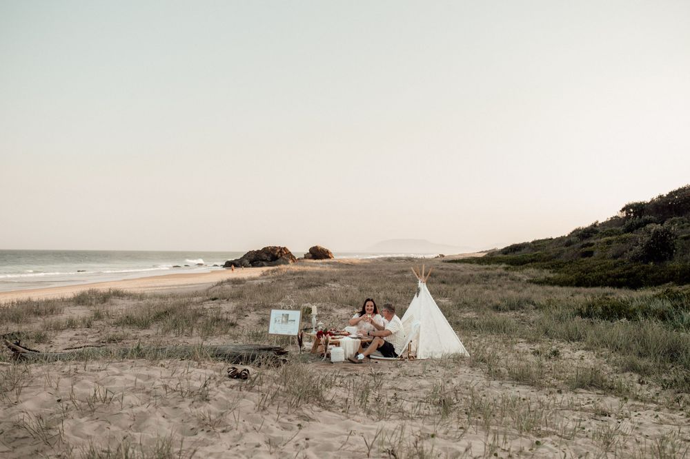 Dreamy coastal landscape with white teepees and rustic decor set up for a beach gathering.