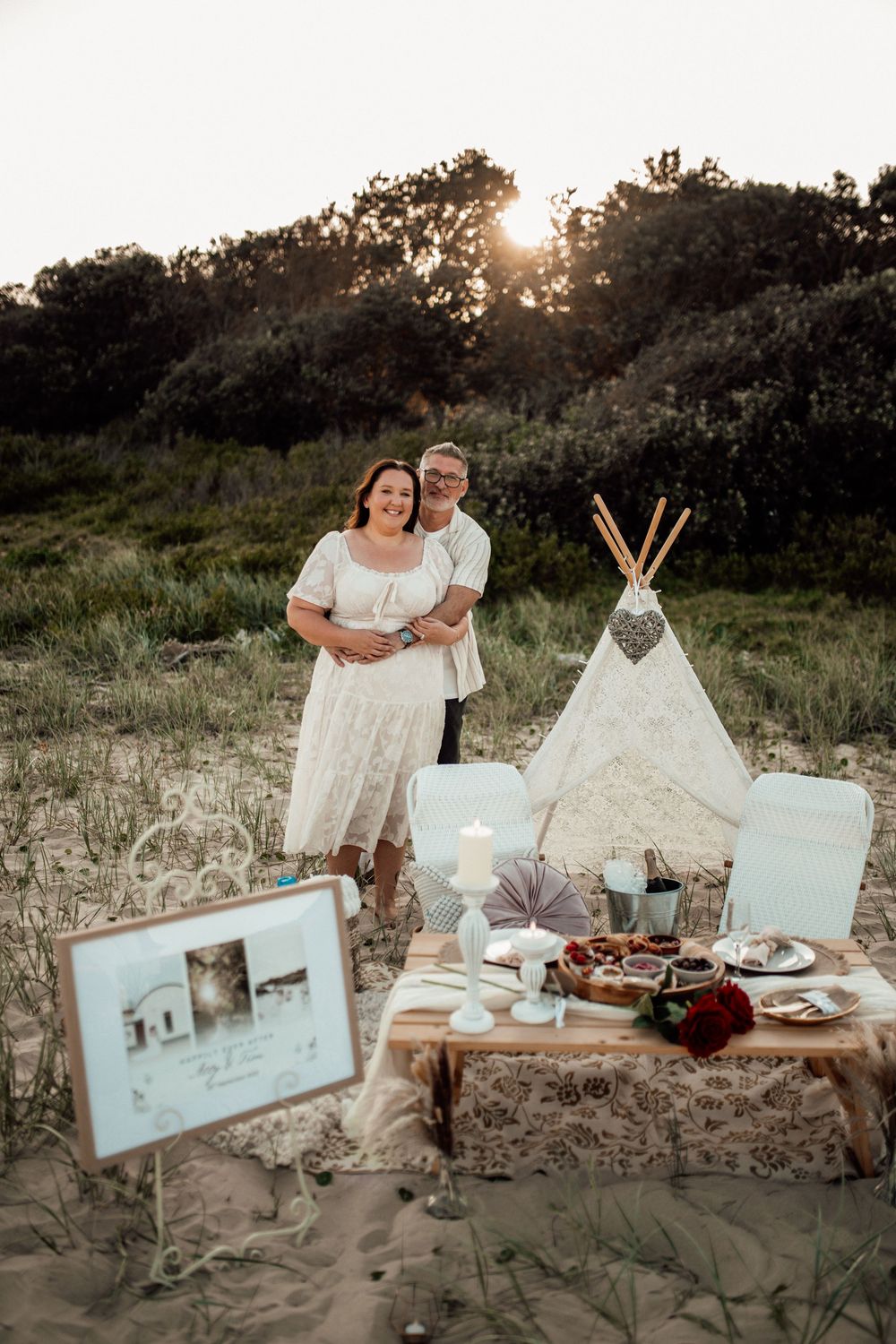 Couple shares intimate outdoor beach picnic with decorative white teepee tents and charcuterie spread at sunset.