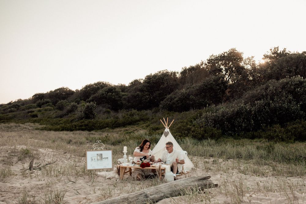 A romantic beach picnic setup with teepees and blankets on sandy dunes at sunset.