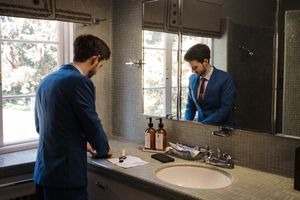 Person in blue shirt looking into bathroom mirror with sink fixture and natural window lighting.