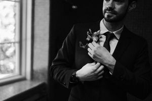 A black and white image shows a suited figure adjusting a floral boutonniere by a window.
