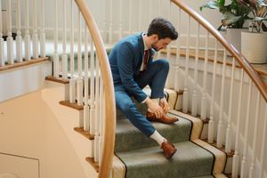Person in a navy suit and brown shoes adjusts laces while sitting on a carpeted staircase with white railings.