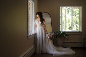A bride in a white flowing wedding dress stands gracefully by a sunlit window in a vintage room interior.