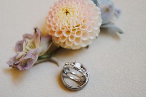 Wedding rings sit nestled among delicate pink and cream dahlia flowers on a light textured background.