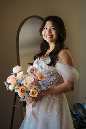 Radiant bride in an off-shoulder lace wedding gown holds a colorful bouquet of pink, peach and blue flowers by a window.