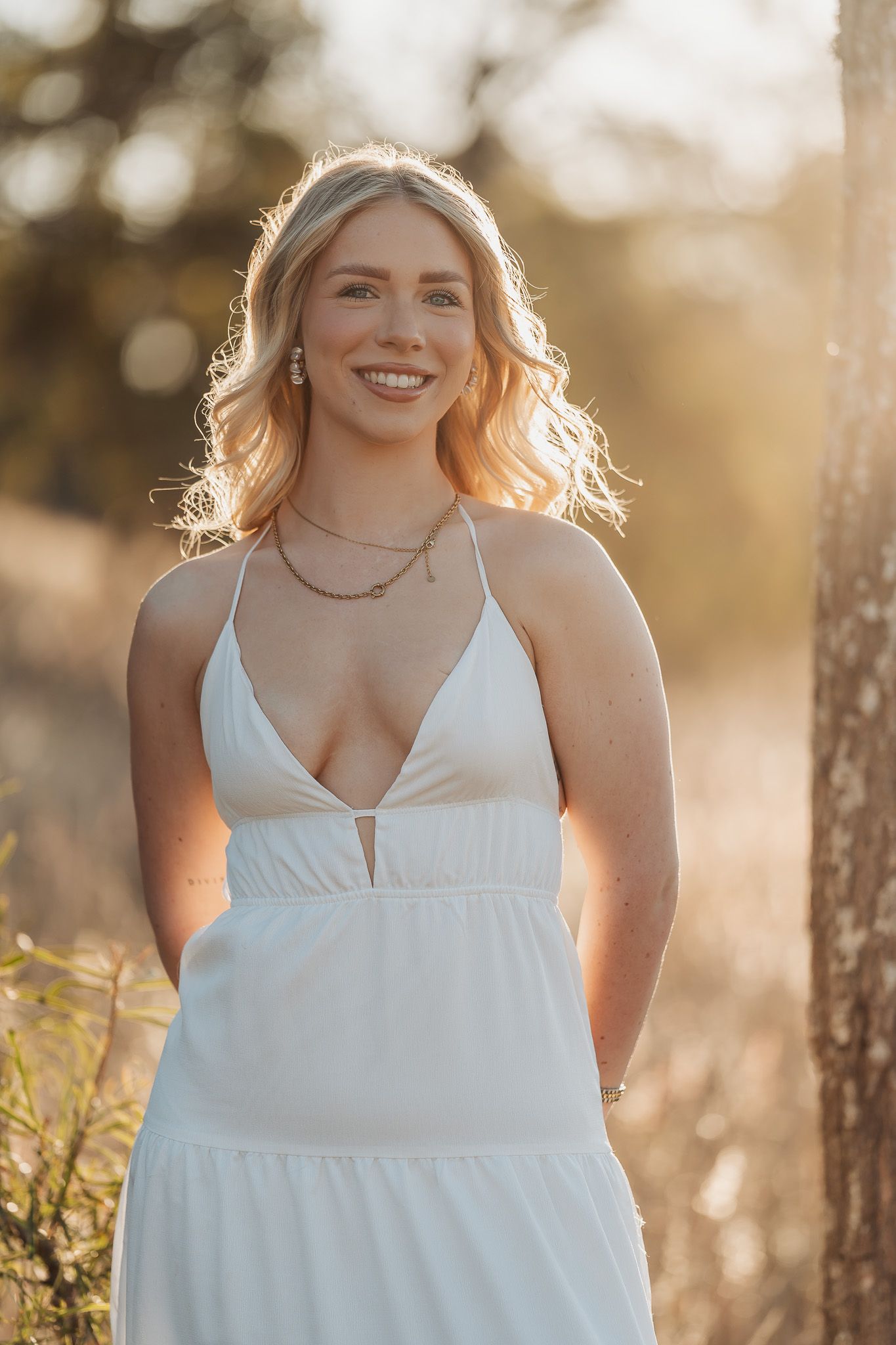 A woman in a white summer dress poses outdoors during golden hour with sunlit hair and a bright smile.