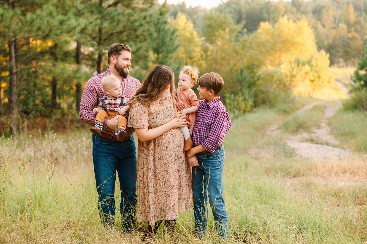 Family stands together on a golden grass hillside during autumn sunset with trees in the background.