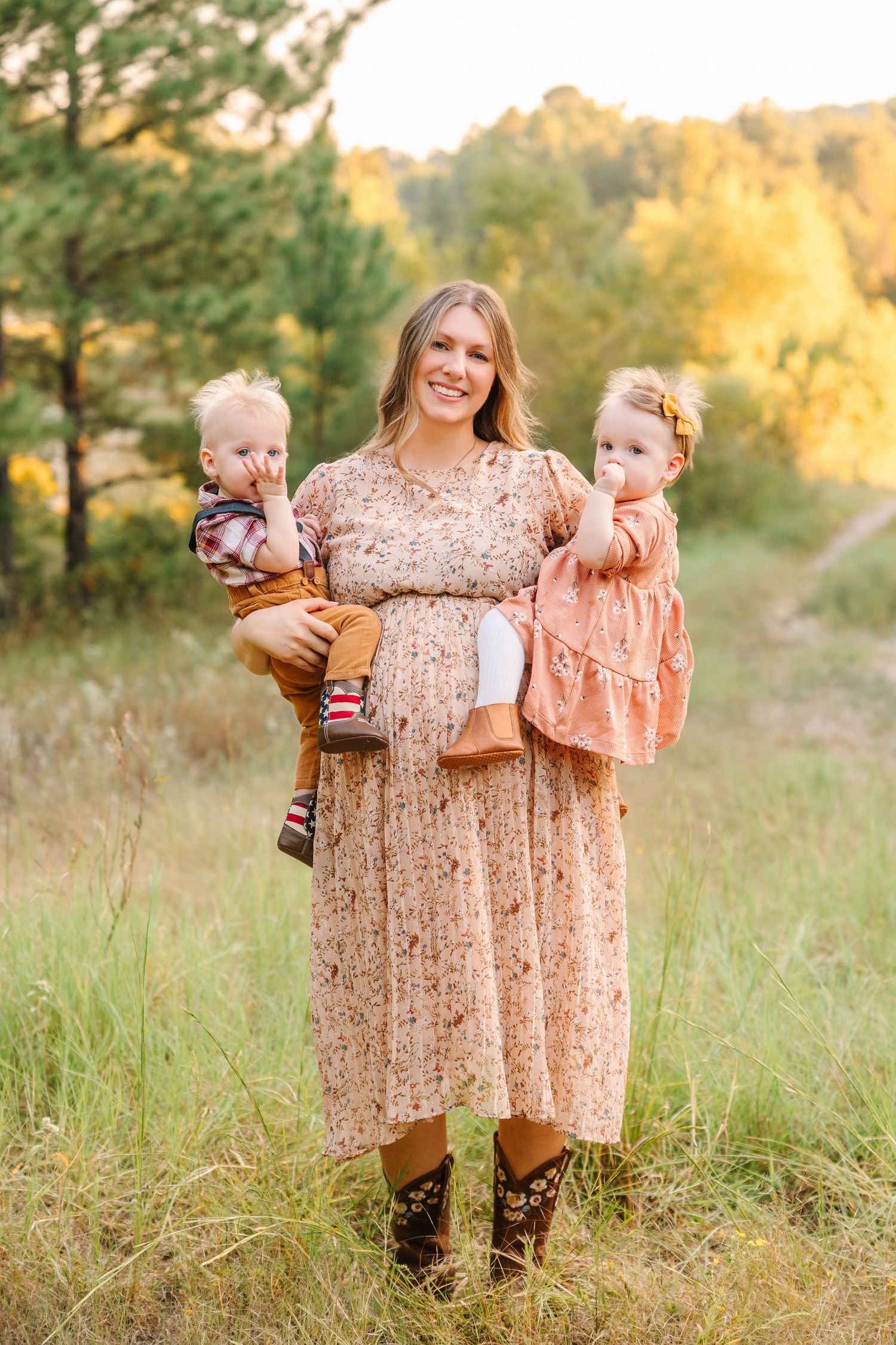 A pregnant woman in a floral dress and cowboy boots stands in a golden field holding two small children.