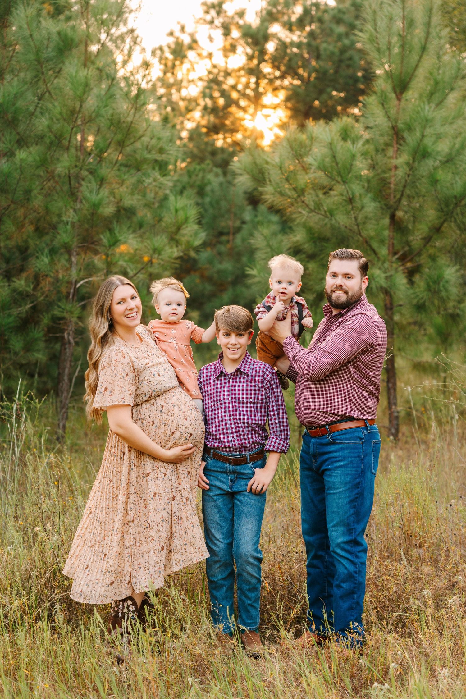 A family stands together playfully posing in a grassy field at sunset with pine trees in the background.