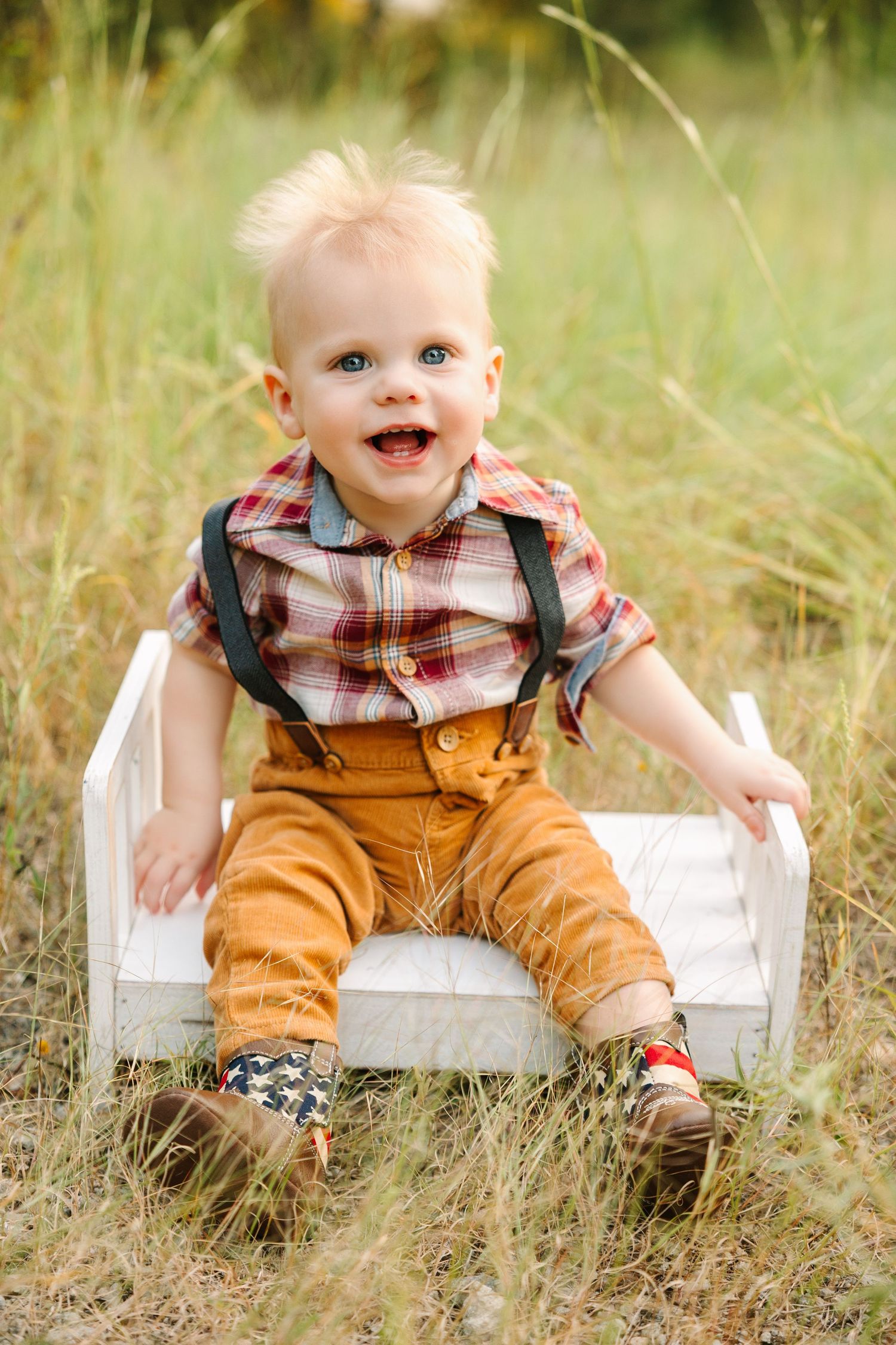 Baby wearing brown overalls sits in white wooden box surrounded by tall grass.