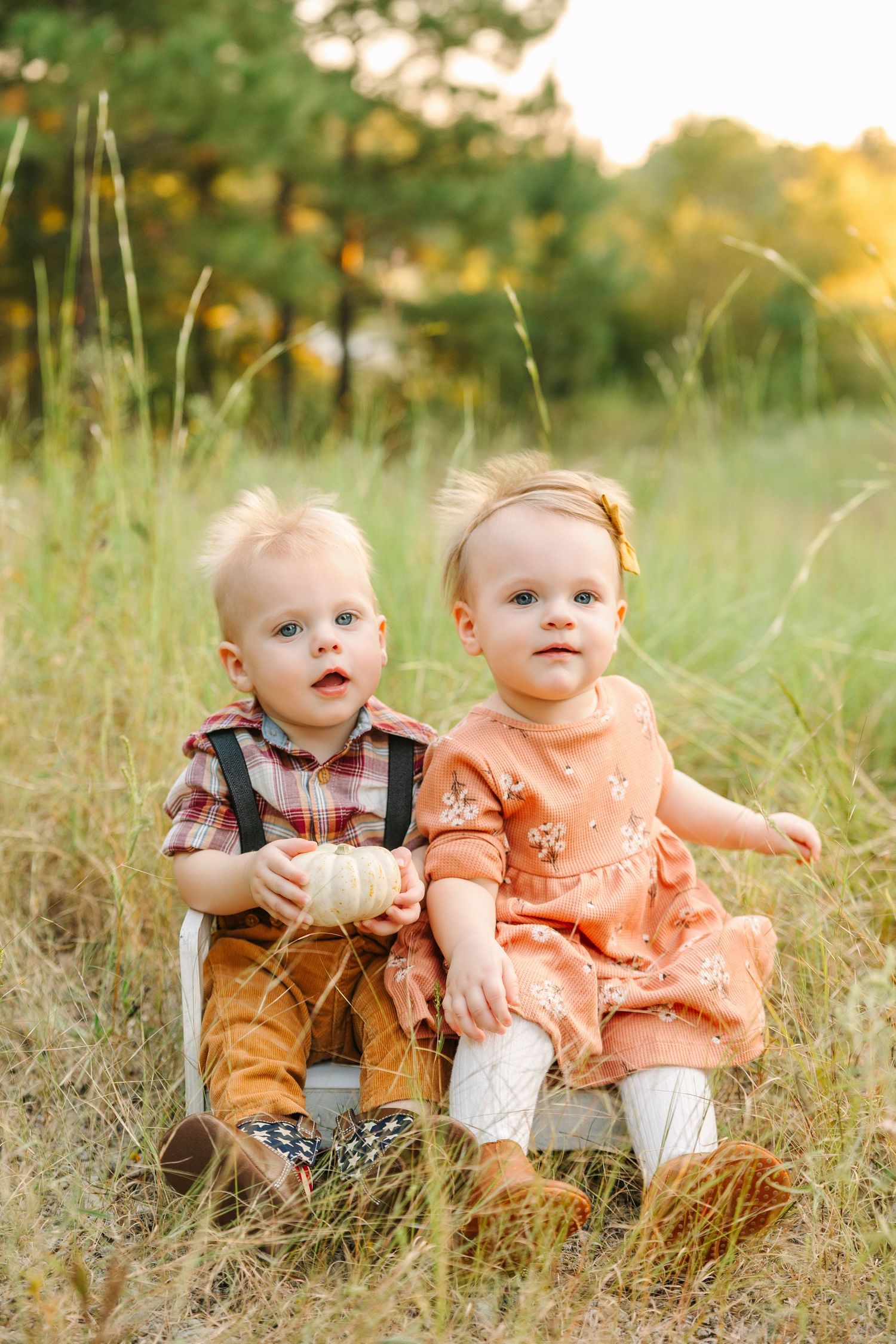 Two small children sit together in a grassy field at golden hour wearing orange and plaid outfits.