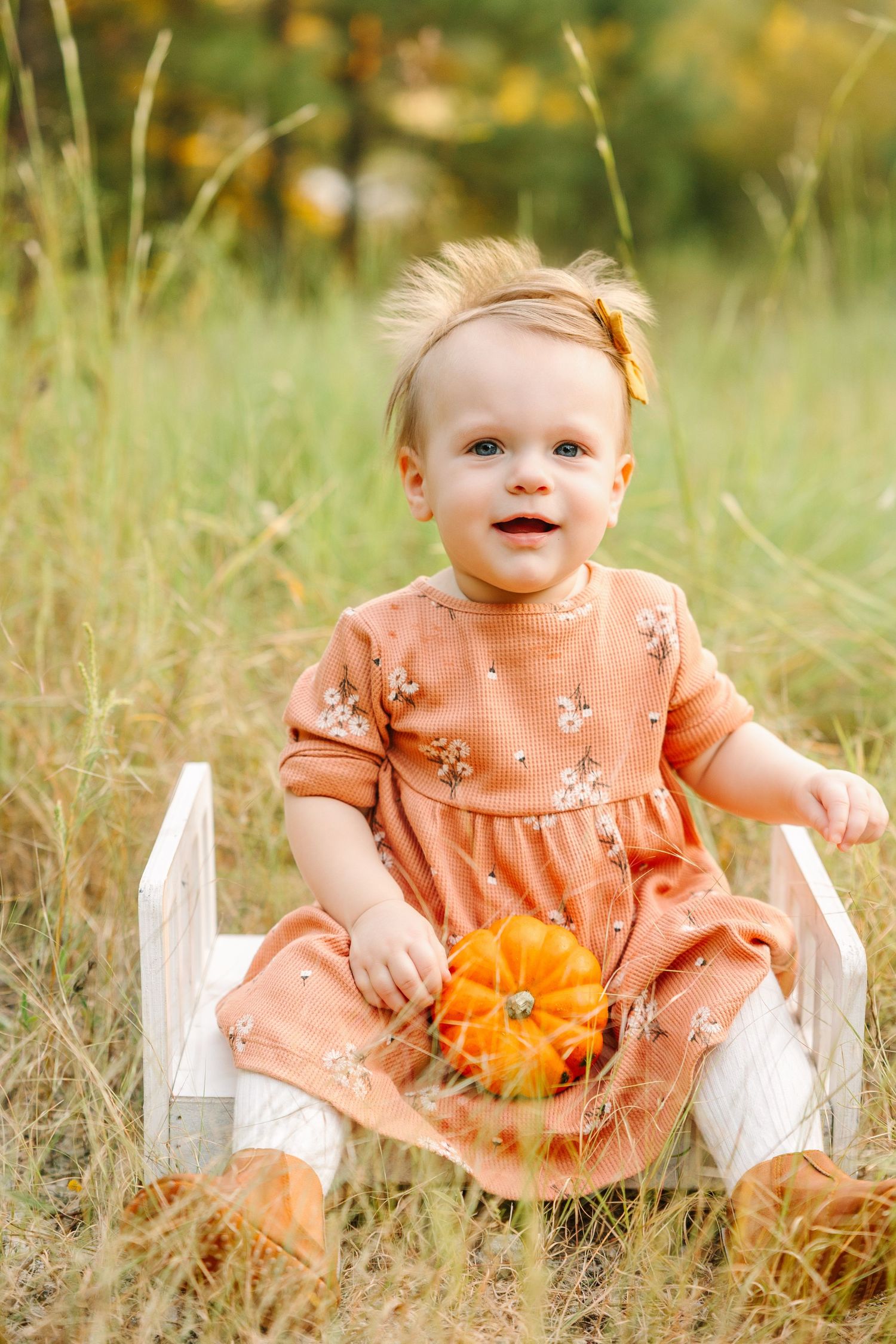 Baby in peach floral dress sitting in white decorative chair with orange flowers in an outdoor field setting.