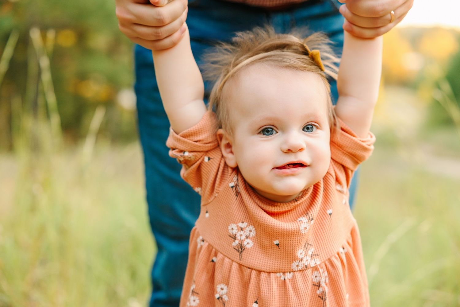 A child wearing a peach-colored dress is held up while standing in a grassy field during golden hour.
