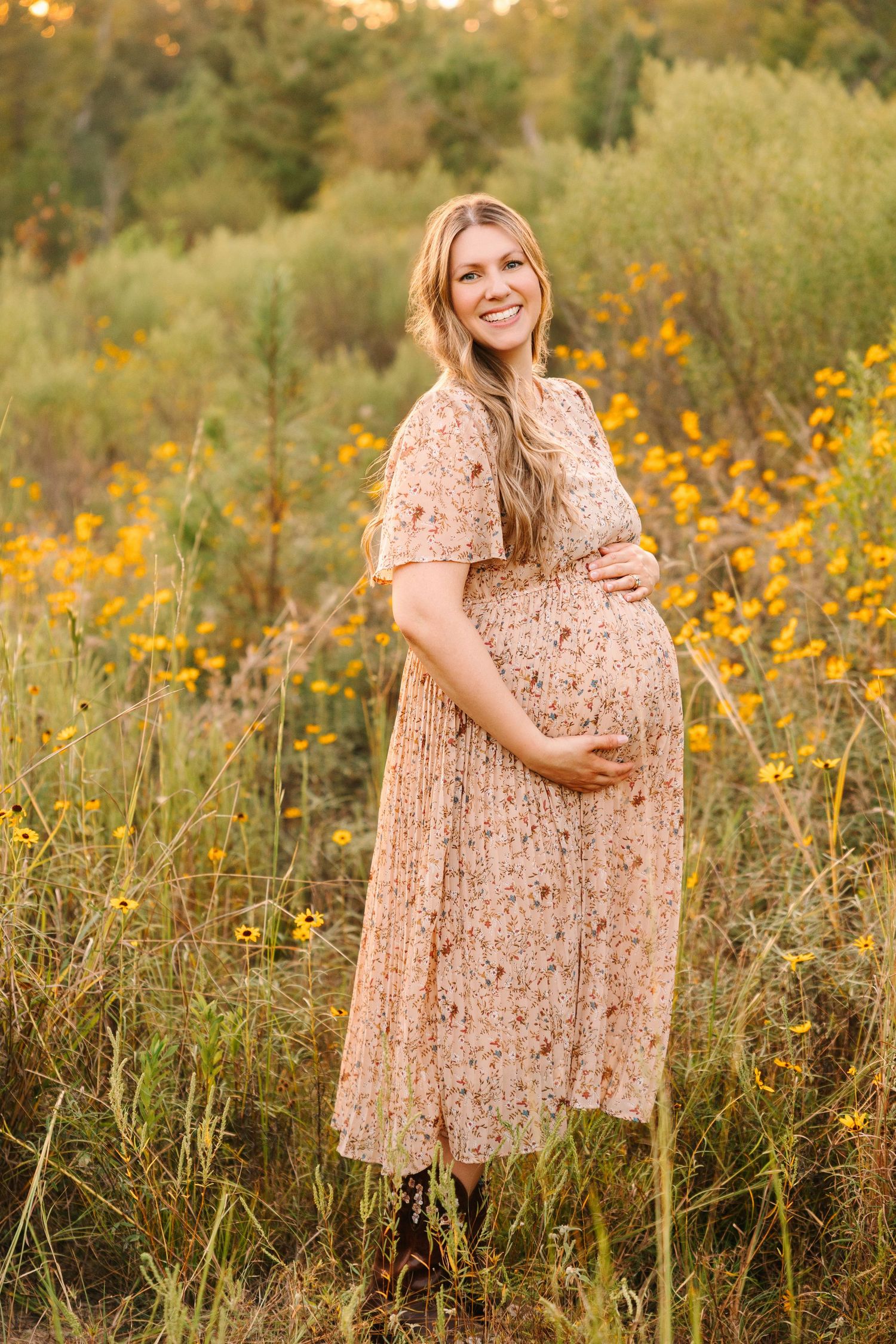 A pregnant woman in a peach floral dress stands in a field of yellow wildflowers during golden hour.