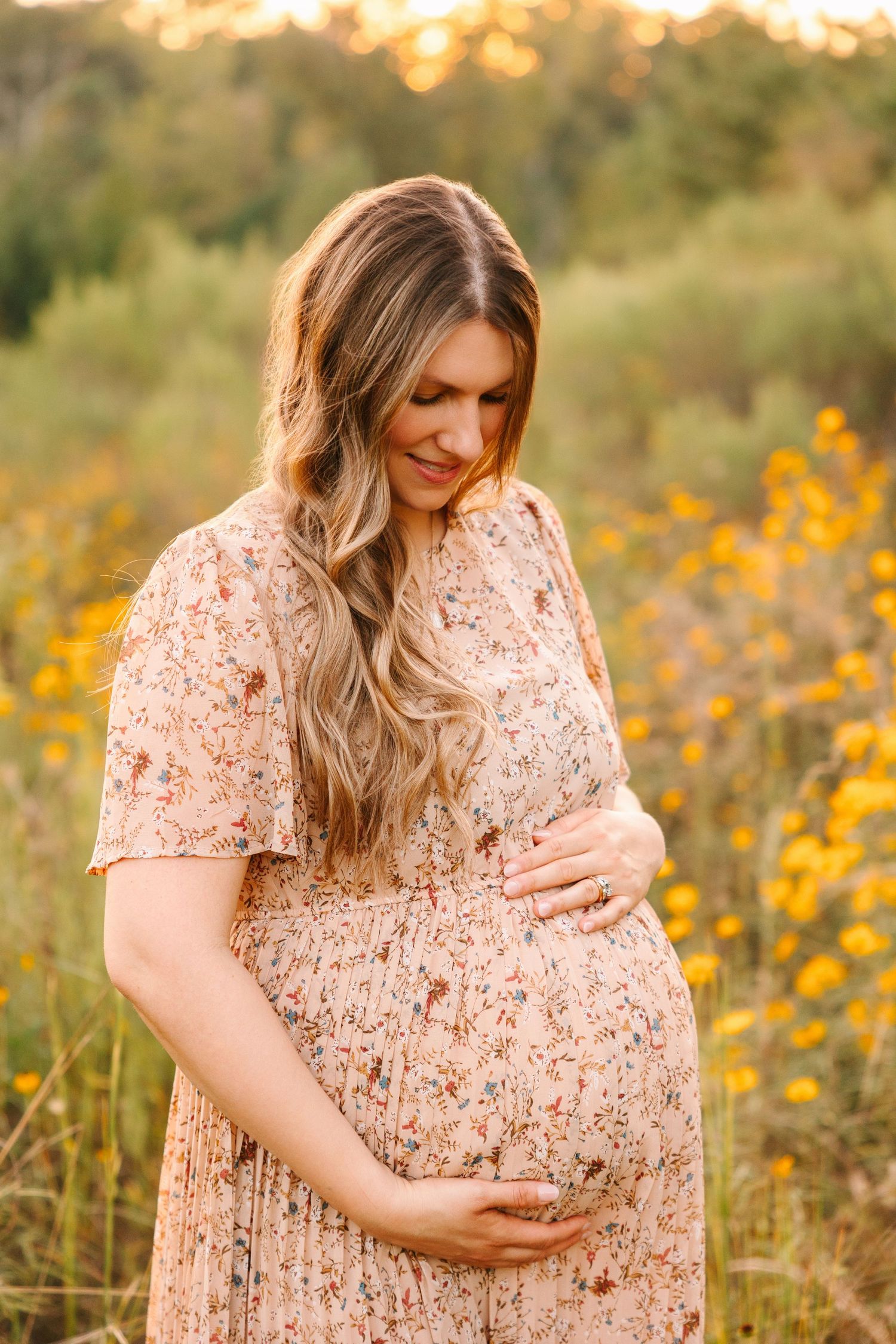 Expecting mother in pink floral dress standing in sunlit wildflower meadow during golden hour sunset.
