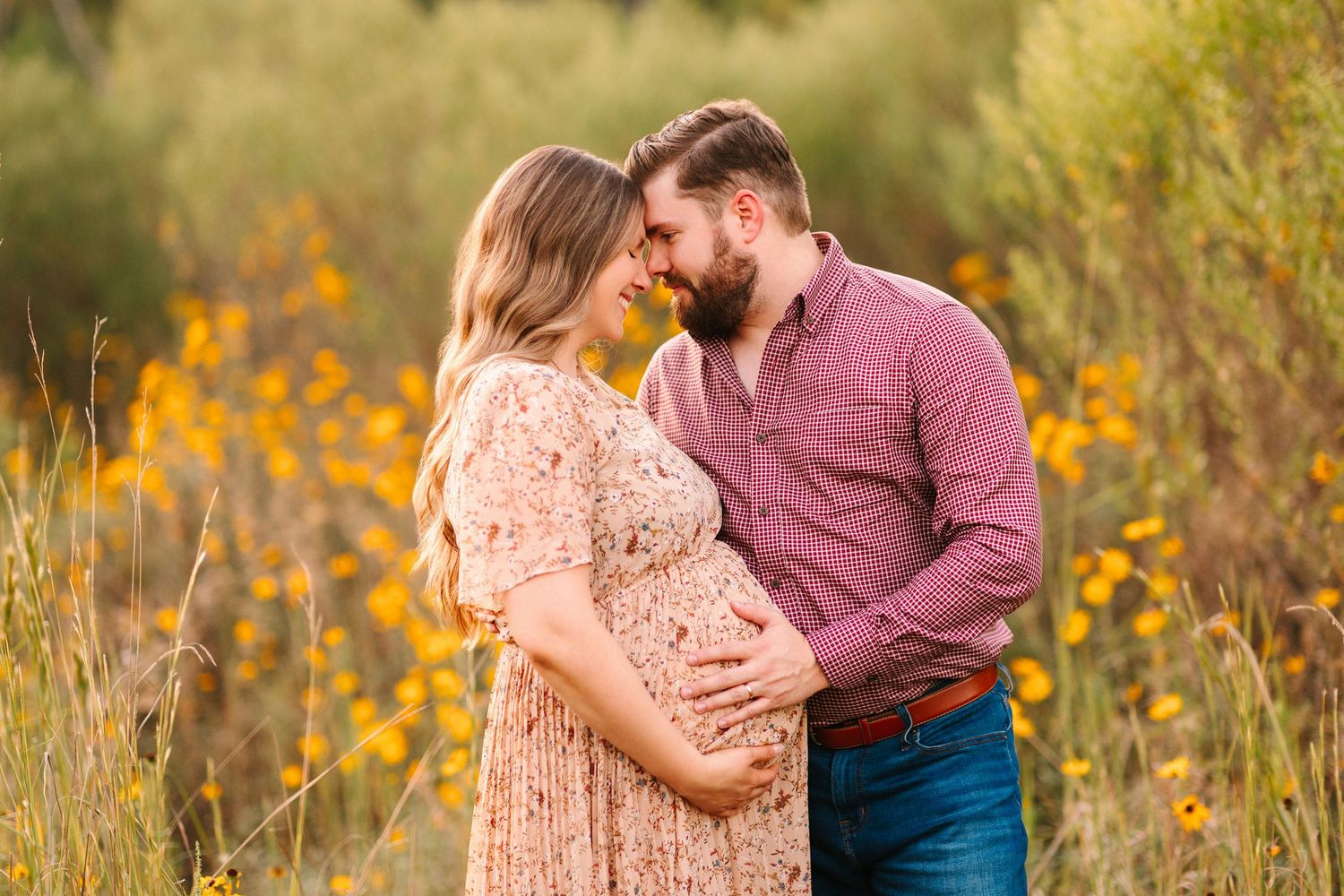 Intimate maternity photo of couple embracing in field with golden wildflowers at sunset.