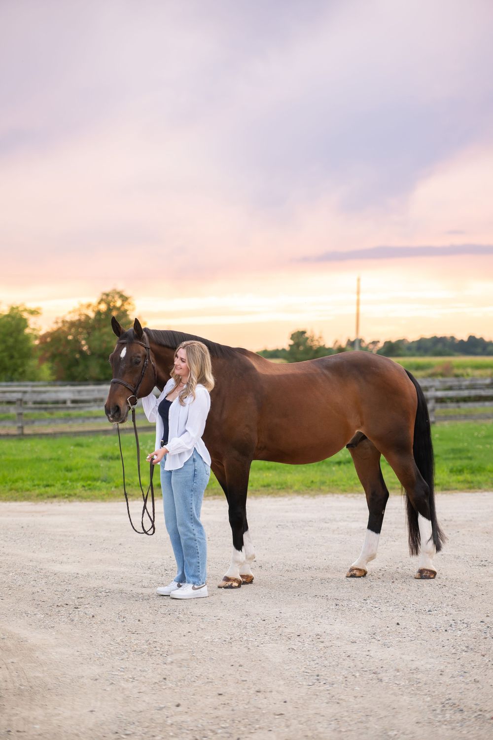 A person in jeans stands beside a brown horse in a gravel paddock during golden sunset light.