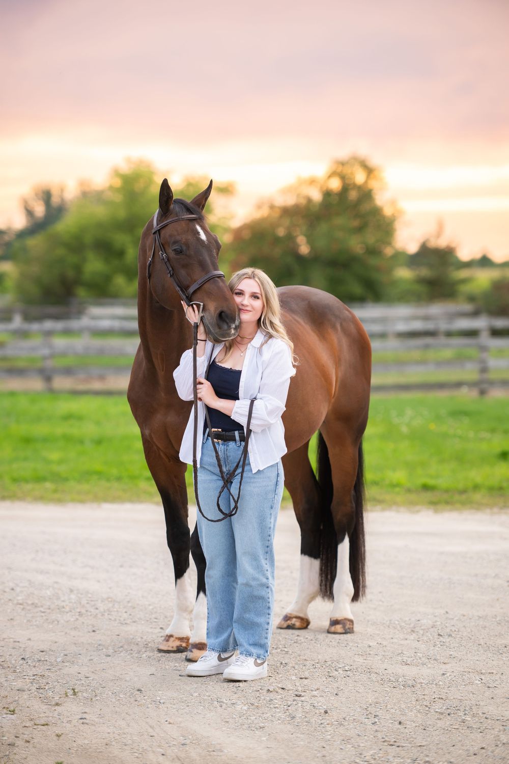 A warm sunset photo shoot with a brown horse and someone in denim overalls and white shirt on a gravel path.