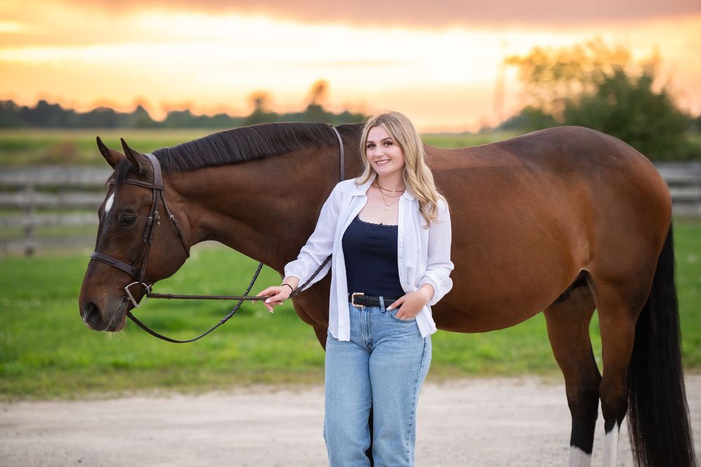 Person stands with brown horse against sunset sky at farm during golden hour photo session.