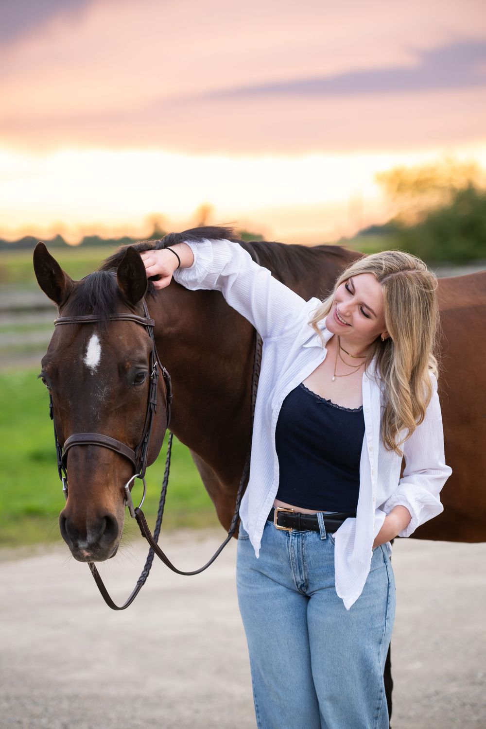 Playful equestrian photo session featuring a dark brown horse and someone in casual denim and white attire at sunset.