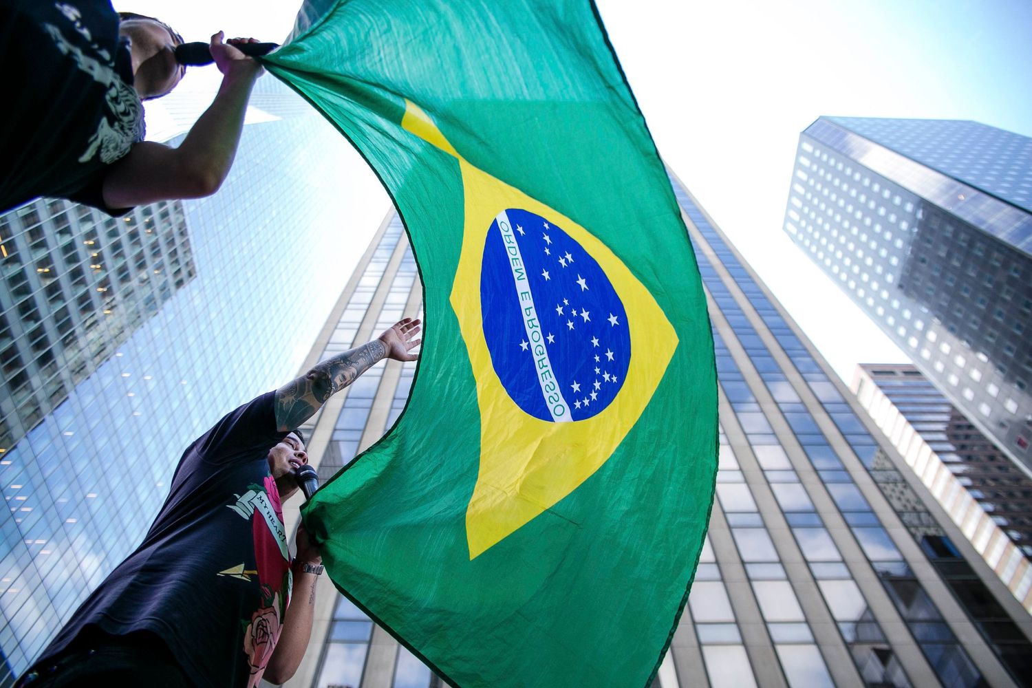A Brazilian flag waves against a backdrop of modern skyscrapers and glass buildings in a financial district.